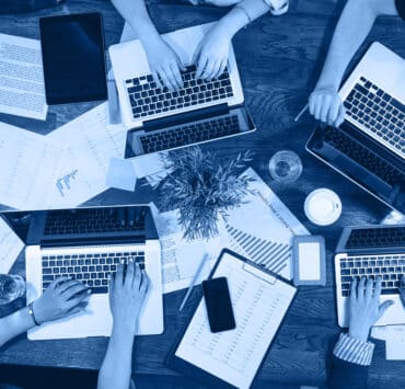 A bird's eye shot of four people working on their laptops, surrounded by business documents.