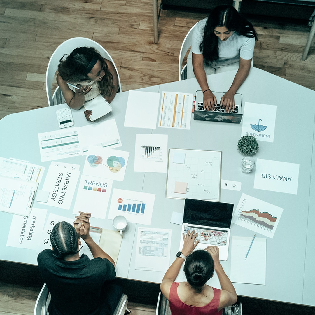 Four women of color sit at a table, working on their computers or on various printed-out finance documents