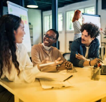 Two women of color and a man sit at a table in conversation with each other with documents laid in front of them