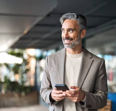 A Latino businessman wearing a taupe blazer smiles and looks off to the side of the camera while holding his iPhone