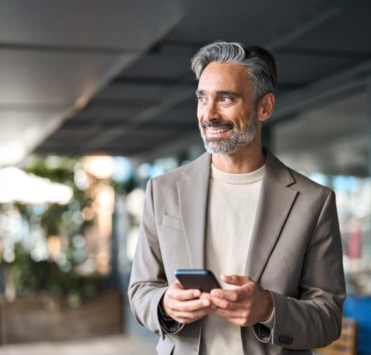 A Latino businessman wearing a taupe blazer smiles and looks off to the side of the camera while holding his iPhone