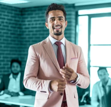 A Latino leader stands and smiles for a photo, wearing a light blush colored blazer and red patterned tie, against a blue-green background