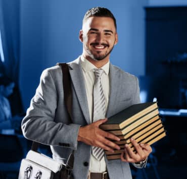 A Latino man holds a stack of books while wearing a gray suit and messenger bag against a blue background